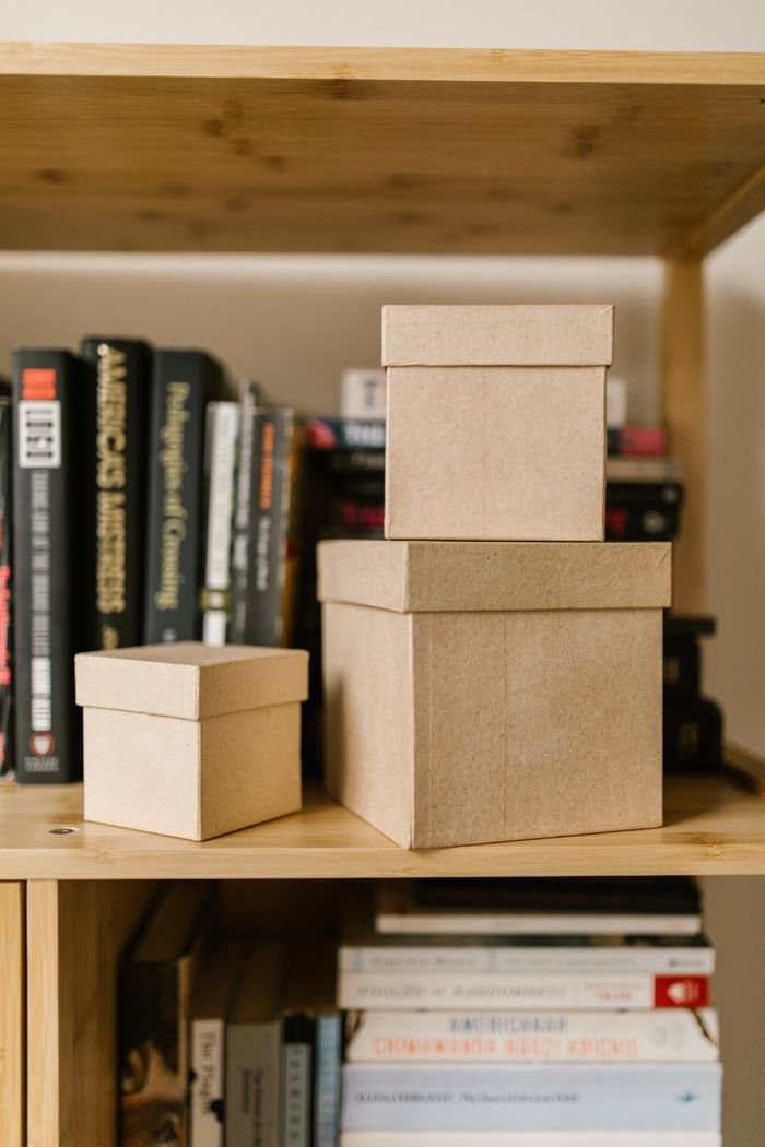 About Close-up of cardboard boxes on a wooden bookshelf surrounded by various books.
